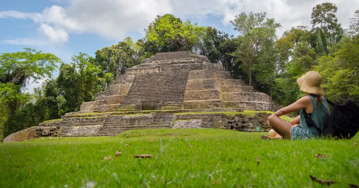 A young woman is sitting in the grass looking at one of the pyramids of the Mayan site of Lamanai