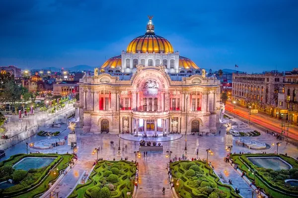 The grand Palacio de Bellas Artes illuminated at twilight in Mexico City, featuring its famous orange dome and surrounding landscaped plaza.