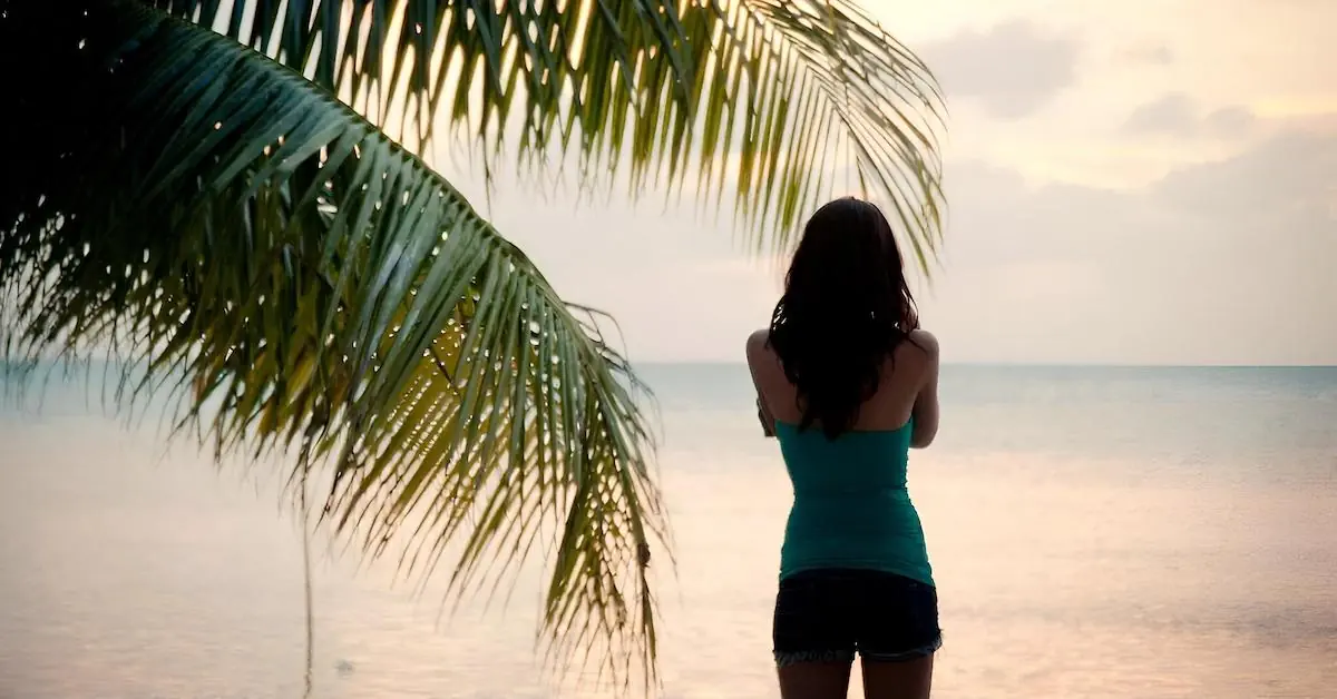A young woman is looking at the Caribbean Sea next to a palm tree during sunset in Caye Caulker