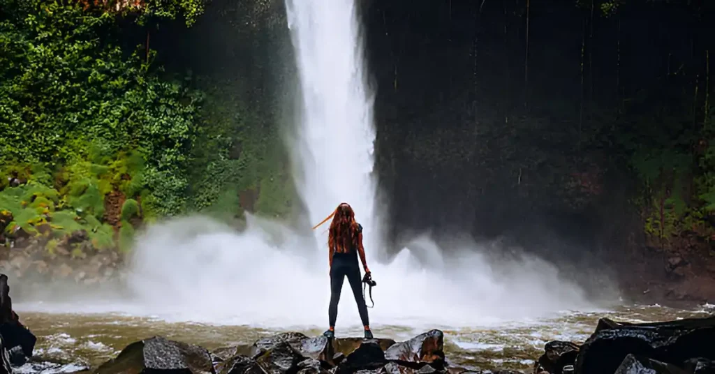 A young woman is standing with a camera in front of the waterfall La Fortuna in Costa Rica