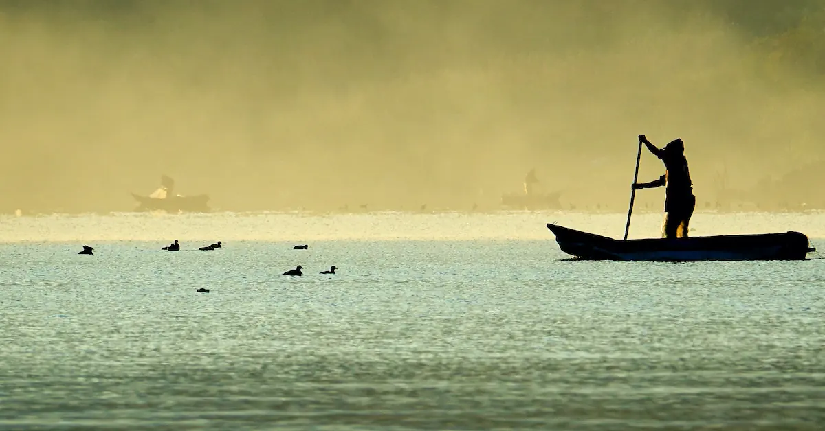 A fisherman is paddling his wooden canoe during sunset in Lake Atitlan while a few ducks are swimming with other fishermen in the background