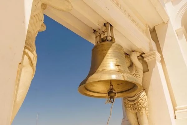 A large bronze bell hanging from the beautiful white colonial architecture of the Leon Cathedral in Nicaragua against a clear blue sky.