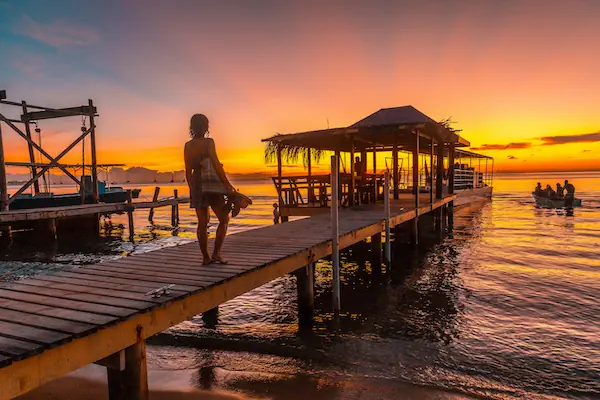 A young woman stands on a wooden dock watching a vibrant sunset over the calm ocean in Roatan, Honduras.