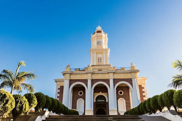 Stone steps lead up to a beautiful historic church with white arches and a yellow bell tower in Santa Ana, El Salvador, set against a clear blue sky.