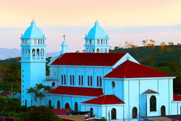 The historic Santa Lucia Church in Juayua, El Salvador, stands out with its bright white facade and distinctive red roofs against a soft evening sky.