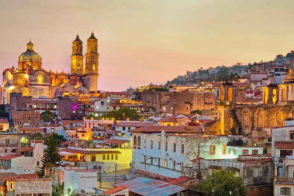 The charming hillside town of Taxco at dusk, showcasing traditional white colonial buildings, red tile roofs, and the illuminated Santa Prisca church.