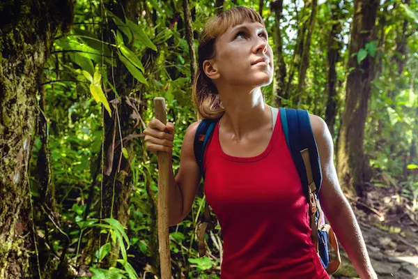 A detailed close-up photograph of a female hiker wearing a red top and backpack, looking upwards while holding a wooden walking stick, on a shaded path within a thick, moss-covered tropical rainforest.