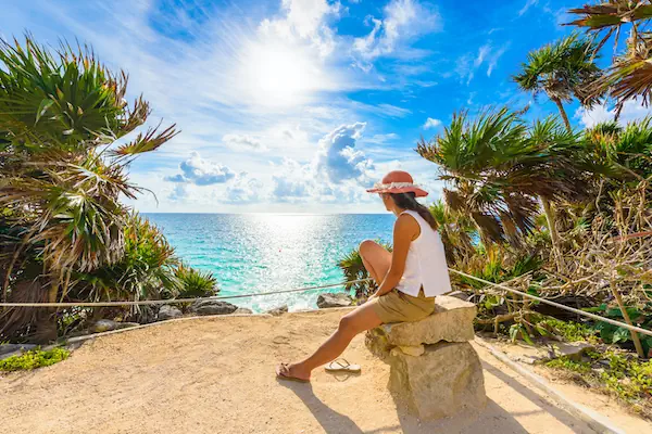 A traveler relaxing on a rocky coastal cliff under palm trees while looking out at the bright turquoise Caribbean Sea in Tulum.