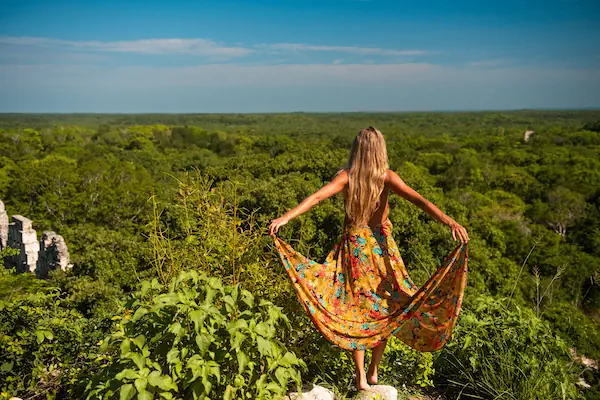 A traveler standing high upon the ancient Uxmal stone ruins looking out over an endless expanse of lush green jungle in Mexico.