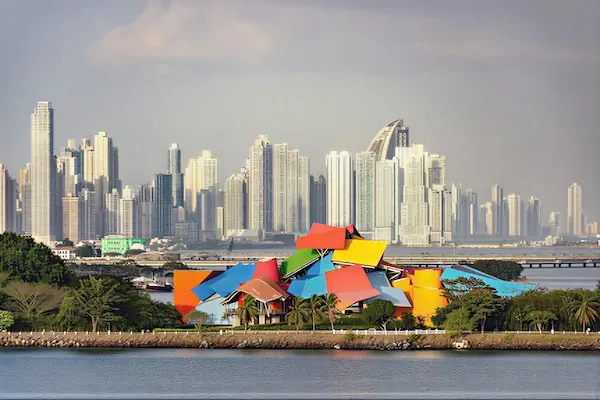 The striking, multi-colored Frank Gehry-designed Biomuseo building sitting on a green peninsula, contrasted against the towering modern skyscrapers of the Panama City skyline across the bay.