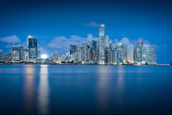 A stunning twilight view of the modern Panama City skyline, with illuminated skyscrapers reflecting beautifully across the calm, deep blue waters of the bay.
