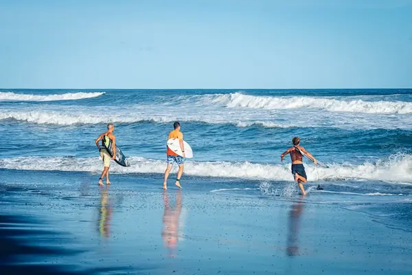 El Paredon, Guatemala Surfers carrying boards into the Pacific waves on the reflective volcanic black sand of El Paredon, Guatemala.