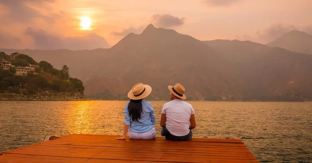 A couple wearing straw hats sits on a wooden dock at sunset, looking across the golden waters of Lake Atitlan toward the Rostro Maya mountain profile in Guatemala.